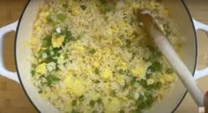 Fried rice with scrambled eggs and scallions being stirred in a white pot on a wooden surface.